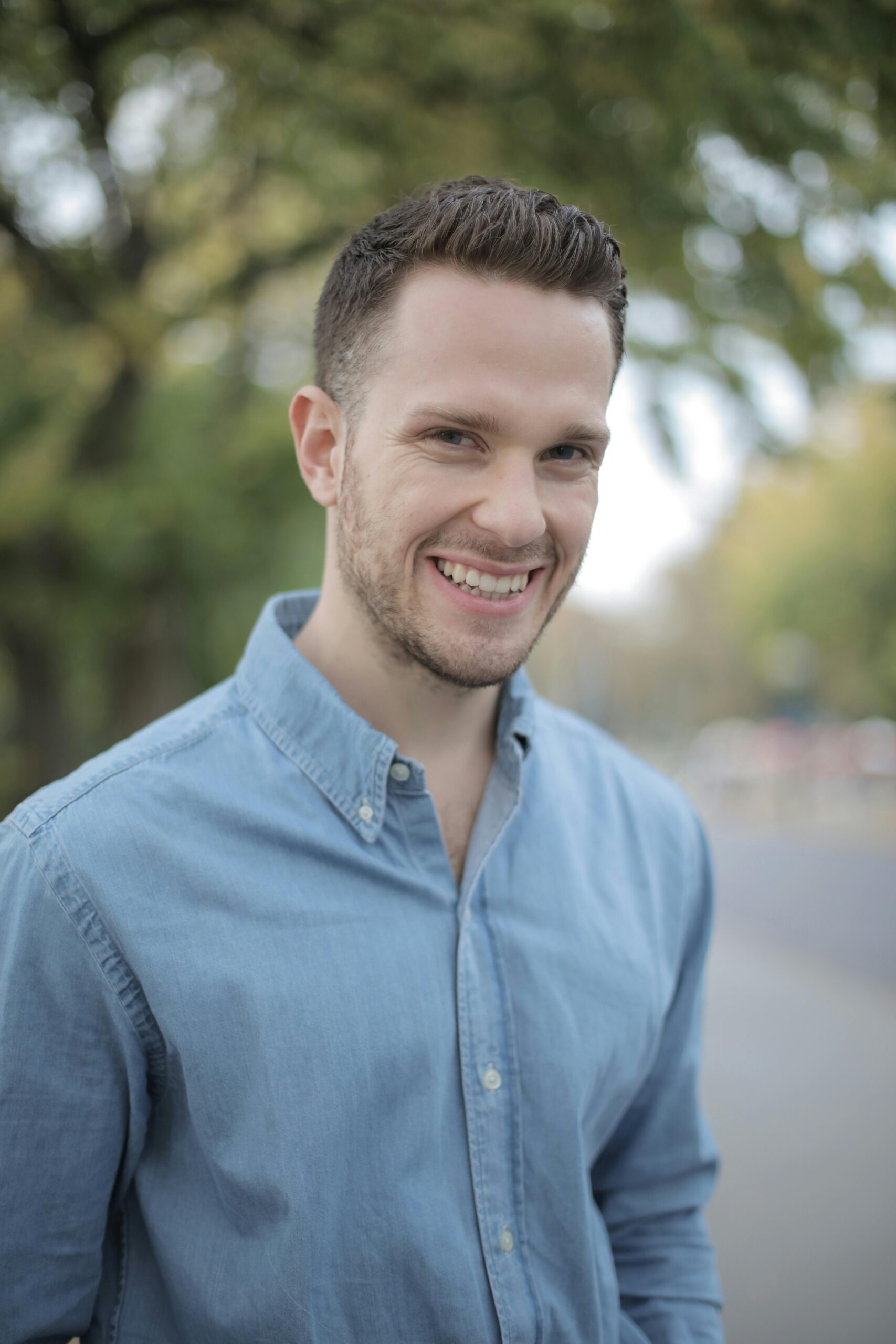 A smiling man in a denim shirt enjoying a sunny day outdoors.