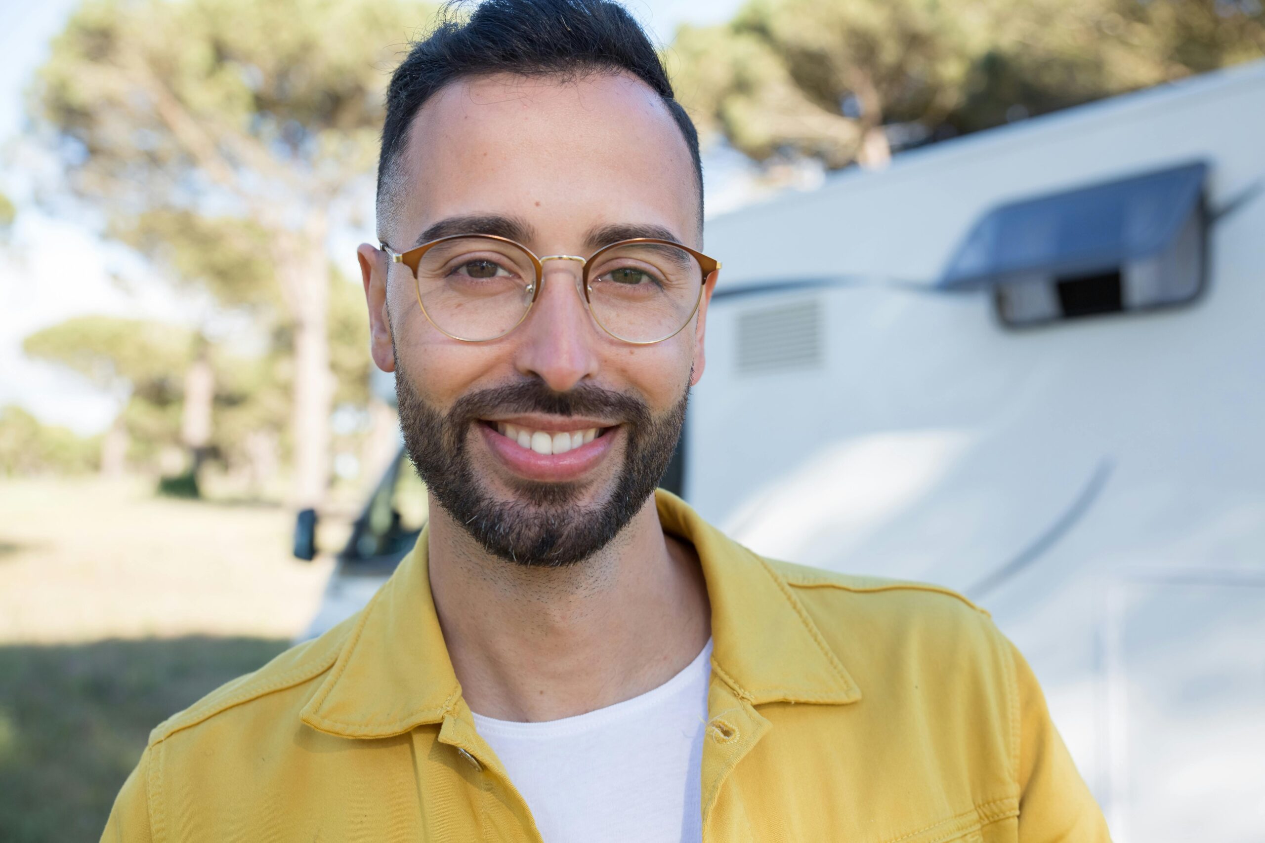 A cheerful bearded man wearing glasses is smiling outdoors in a natural setting.